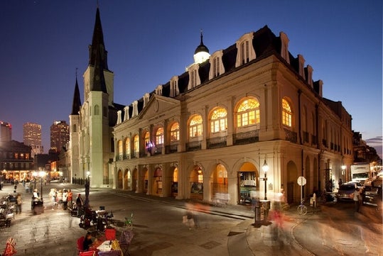 Presbytère Louisiana State Museum, a large building with many windows. There is a street in front of the building with cars parked along it. It is night time and the building is lit up.