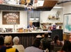 A woman working in a kitchen setting, conducting a cooking demonstration. The surroundings include a countertop, table, and menu, typical of a restaurant or cooking school environment.