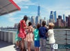A group of children accompanied by a woman enjoy the view of New York City's skyscrapers from aboard a ferry on the water.