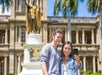 A man and a woman standing in front of a building, smiling and taking a picture together outdoors with a clear sky and a tree in the background as part of an activity called Shaka Guide: Driving and Walking Audio Tours.