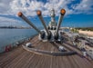 View from the deck of a battleship showing large artillery guns pointing forward, with a harbor and cityscape in the background under a partly cloudy sky.