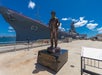A statue of a naval officer on a black pedestal is in front of a large battleship labeled "63" docked at a harbor under a blue sky.