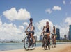 Group of people riding bicycles along a waterfront path with city buildings and cloudy sky in the background.