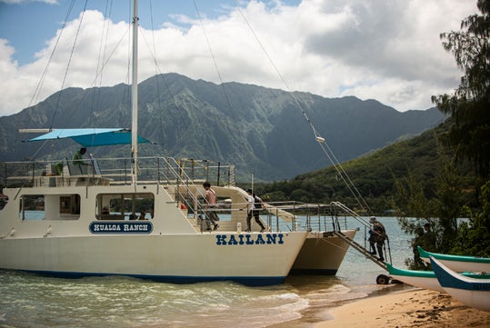 A white boat named "Kailani" is docked at a sandy beach surrounded by lush green mountains. People are boarding the boat using a ramp.