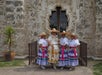 Four women in traditional colorful Mexican dresses and hats stand in front of a historic stone building with an ornate window.