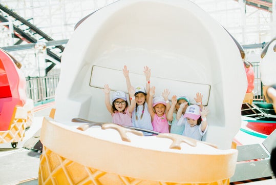 Five children smiling and raising their hands while seated inside an ice cream-themed amusement park ride.