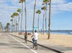 Two people ride bicycles along a beachside path lined with palm trees, with a pier and ocean in the background under a partly cloudy sky.