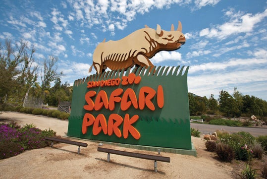 Sign for San Diego Zoo Safari Park featuring a rhinoceros sculpture, surrounded by greenery and benches under a partly cloudy sky.