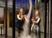 Two women observe and interact with a vertical swirling column of mist in an indoor setting.