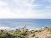 A coastal view with a long pier extending into the blue ocean, surrounded by greenery and scattered palm trees under a partly cloudy sky.