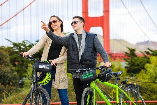 Two people stand with bikes in front of the Golden Gate Bridge; one is pointing ahead while both are smiling and wearing sunglasses.