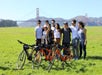 A group of eight people stand on grass beside bicycles, with the Golden Gate Bridge and hills visible in the background on a sunny day.