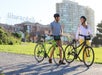 Two people wearing helmets stand with bicycles in a park near the Ghirardelli building, with tall buildings and grass visible in the background.