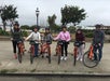 Six people in helmets stand with orange rental bikes on a paved path in a park, with trees and a ship visible in the background on a cloudy day.