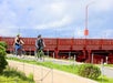 Two people ride bicycles on a paved path near a red bridge on a partly cloudy day.