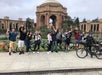 A group of people with bikes pose and wave in front of the Palace of Fine Arts in San Francisco on a cloudy day.