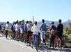 A group of people with bicycles, many wearing helmets, stand on a paved path looking toward the Golden Gate Bridge in the background on a clear day.