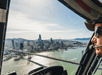 Helicopter pilot wearing sunglasses and headset, flying over a cityscape with skyscrapers and a prominent suspension bridge over green water.