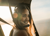 Person wearing a headset, smiling, and looking out the window of an aircraft with sunlight in the background.