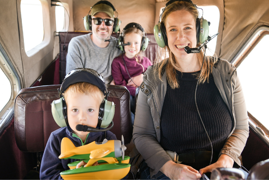 Two adults and two children wearing headsets sit inside a small airplane; one child holds a yellow toy plane.
