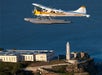 A white and yellow seaplane flies over Alcatraz Island, with the prison building and lighthouse visible below, surrounded by blue water.