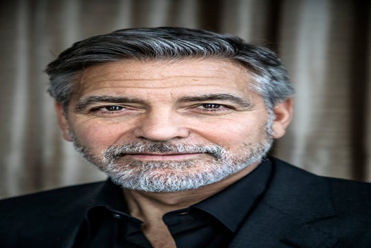A man with gray hair and beard wearing a black jacket and shirt, looking directly at the camera against a blurred curtain background.