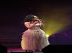 Person singing on stage, wearing a strapless, layered tulle gown, holding a microphone, with stage lights in the background.