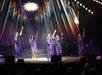 A group of five performers in purple robes sing and raise their hands on a brightly lit stage with a colorful, starry backdrop.