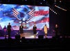 Five performers in uniform sing on stage with an American flag and eagle displayed on a screen behind them.