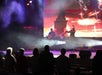 Performer singing on a stage with a large screen displaying fireworks. Audience members are watching.