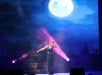 A singer performs on stage under a large moon backdrop, illuminated by purple and blue spotlights.