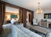Elegant hotel suite with a cozy living area, plush sofa, antler chandelier, and a separate bedroom featuring a green tufted headboard.