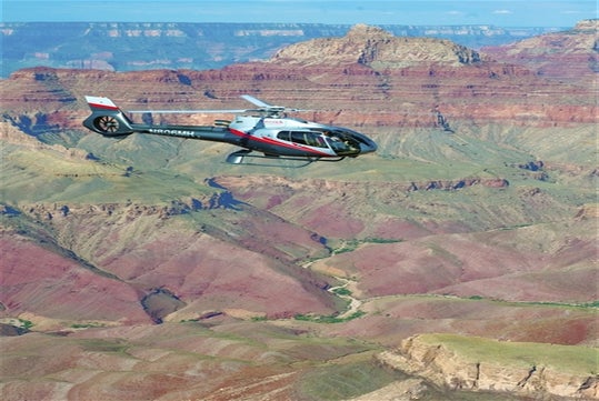 A helicopter flies over the colorful layered rock formations and deep canyons of the Grand Canyon under a clear sky.