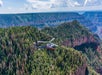 A helicopter flies above a dense forest near the edge of a large canyon with layered rock formations under a partly cloudy sky.