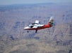 A small red and white airplane flies over the Grand Canyon on a clear day, with layered rock formations visible below.