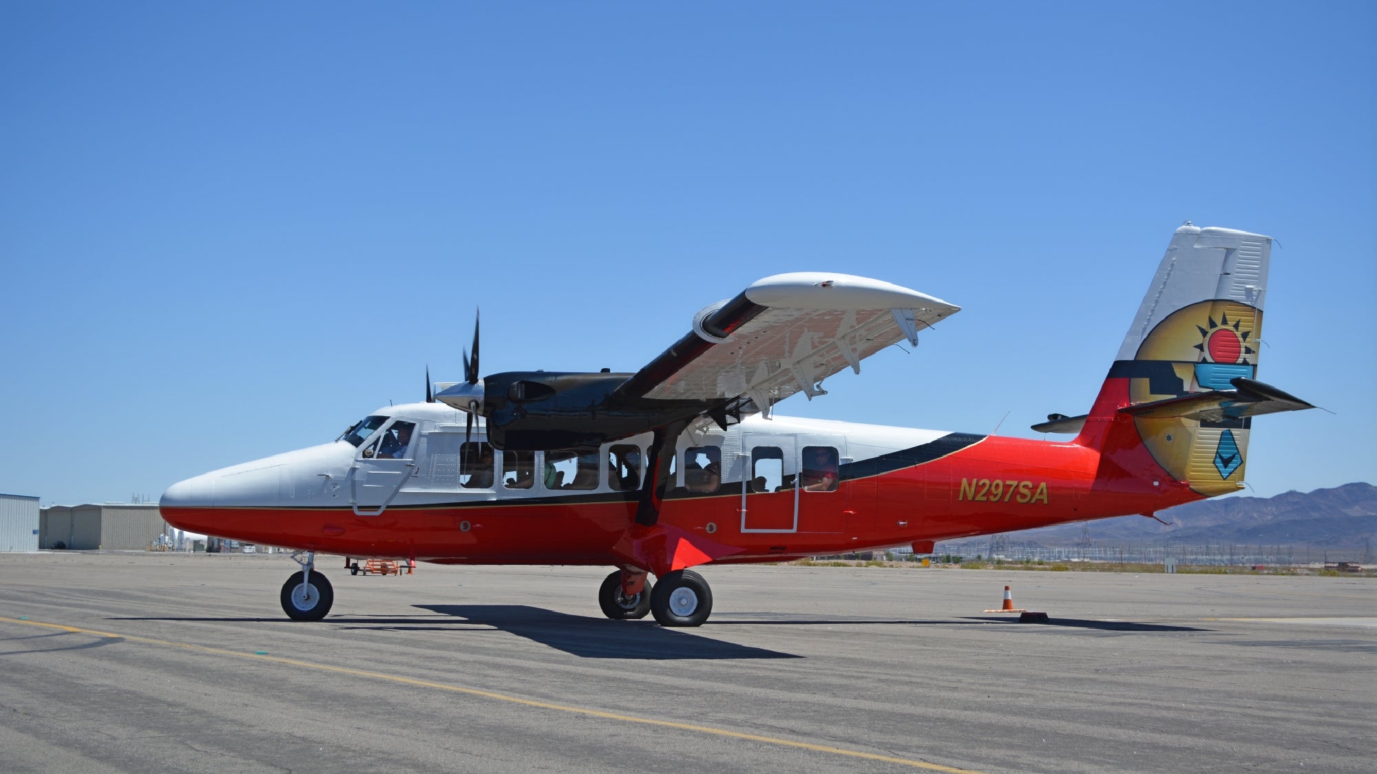 A red and white twin-engine airplane is parked on the tarmac under a clear blue sky, with mountains visible in the background.