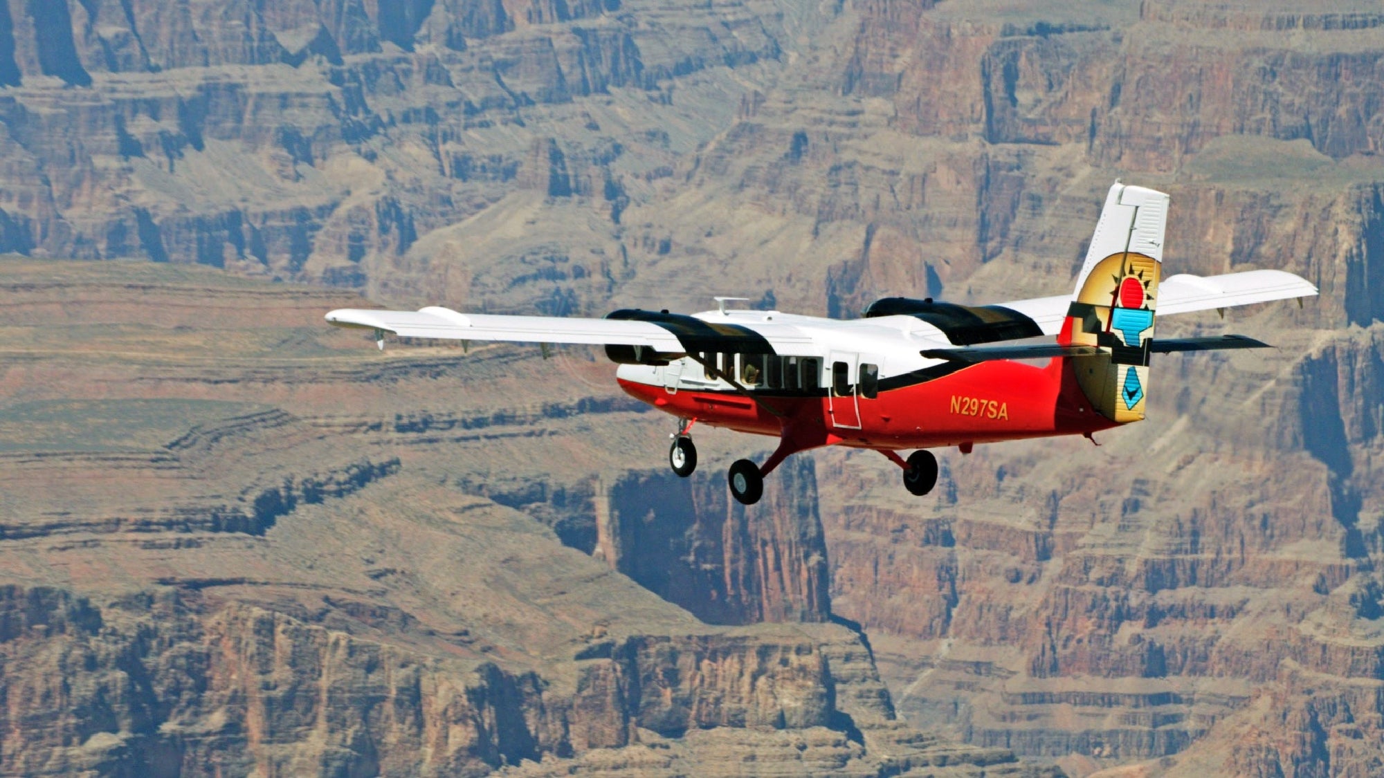 A small red and white sightseeing airplane flies above the Grand Canyon with rock formations visible below.