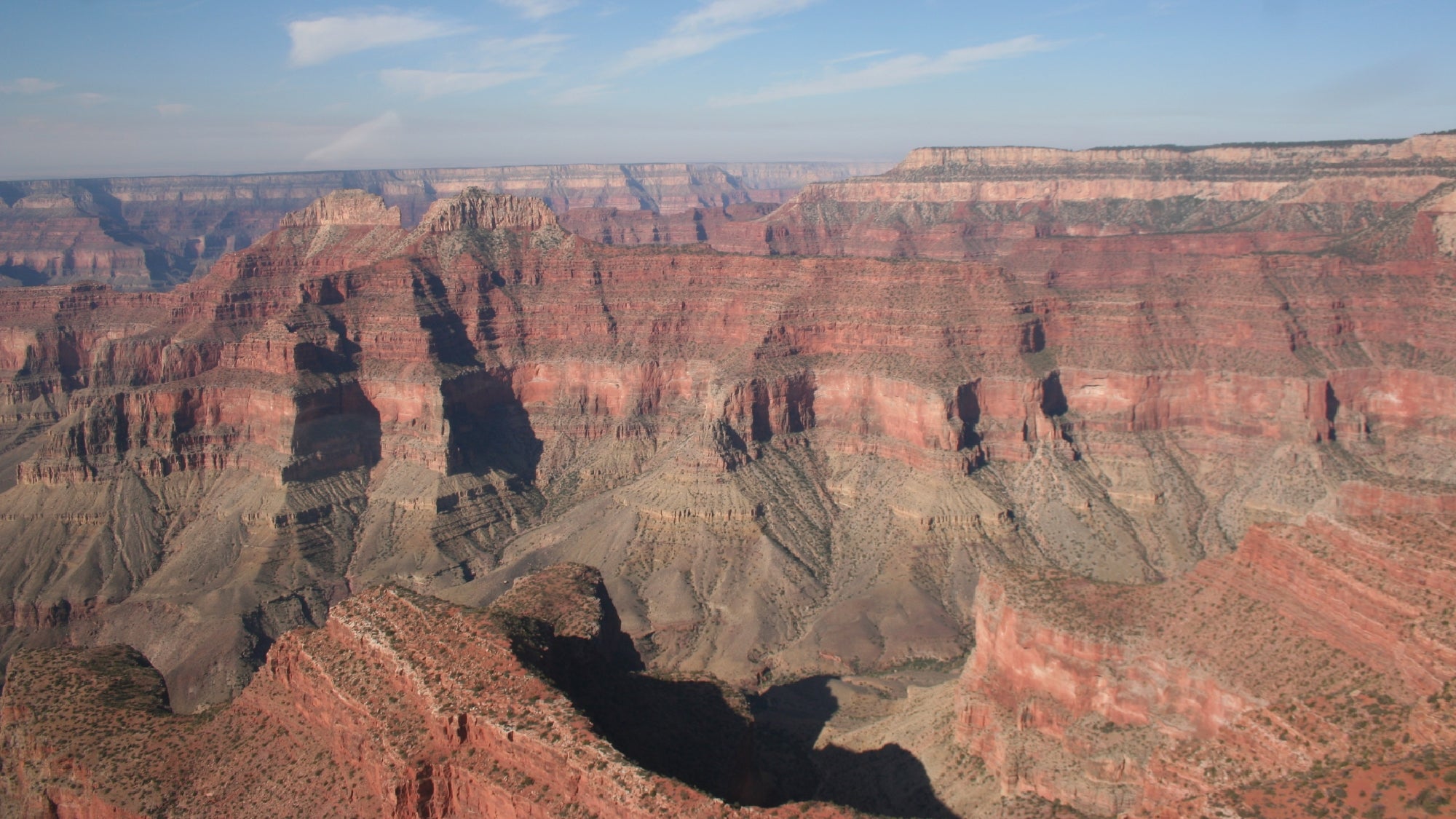 A wide view of the Grand Canyon shows layers of red and brown rock formations under a blue sky with light clouds.