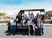 A group of people, including adults and children, pose in front of a black open-air tour vehicle labeled "Buck Wild Grand Canyon Hummer Tours" with canyon scenery in the background.