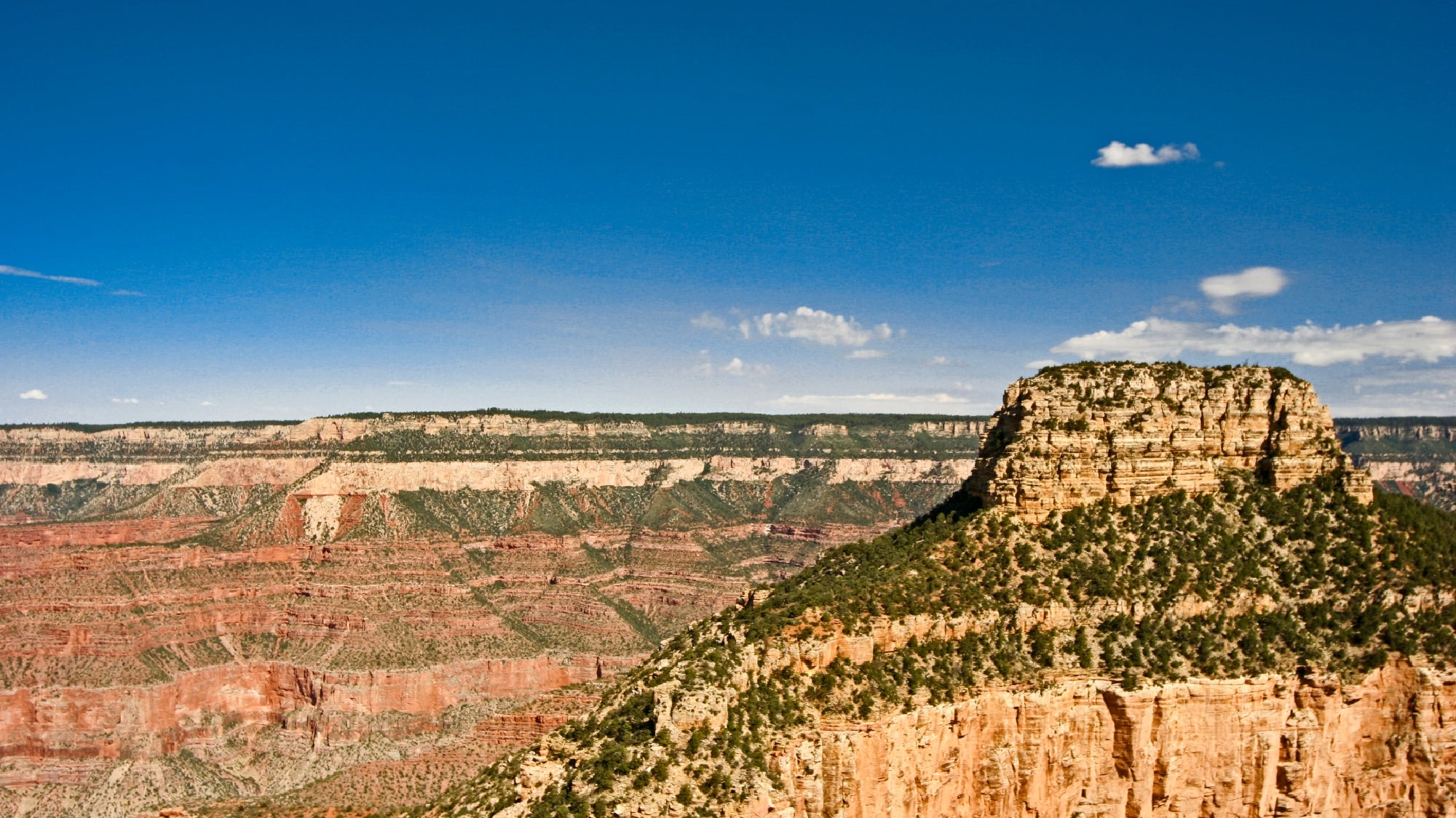 Flat-topped mesa rises above layered red rock cliffs in a canyon landscape under a clear blue sky.