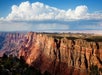 A panoramic view of the Grand Canyon shows layered red rock cliffs under a blue sky with large white clouds and distant rain.