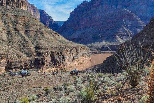 Two Maverick helicopters in the canyon next to the red brown water of Lake Mead with tourists taking photos of the reservoir.