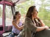 Two women ride in an open vehicle, looking out and smiling as they travel along a road with trees visible through the window.