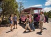 Five people stand near a bright pink open-air jeep parked on a dirt roadside, surrounded by trees and under a blue sky.