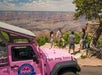 A group of people stands near the edge of a canyon, next to a pink jeep labeled "Pink Jeep Tours," overlooking a scenic landscape.
