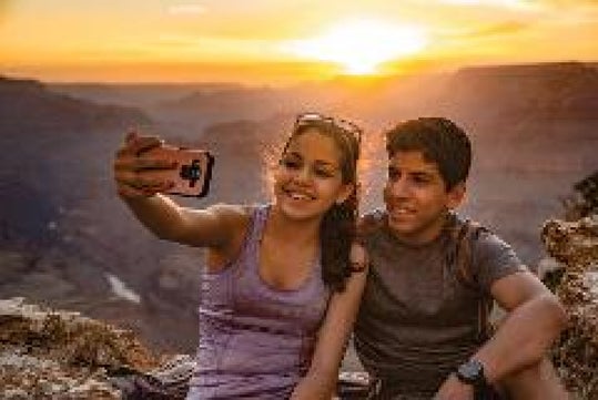 Two people sit on a rocky ledge taking a selfie at sunset, with a canyon landscape visible in the background.