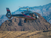 A gray helicopter with red stripes flies over a rugged, mountainous desert landscape.