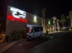 A building with a lit "Maverick" sign and helicopter graphic at night, with a shuttle bus parked in front and palm trees along the entrance.