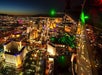 Aerial night view of a brightly lit city with numerous buildings, neon lights, and busy streets, with part of a helicopter visible in the foreground.