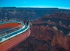A group of people stand on a curved glass skywalk extending over the edge of the Grand Canyon, with steep canyon walls in the background under a clear blue sky.